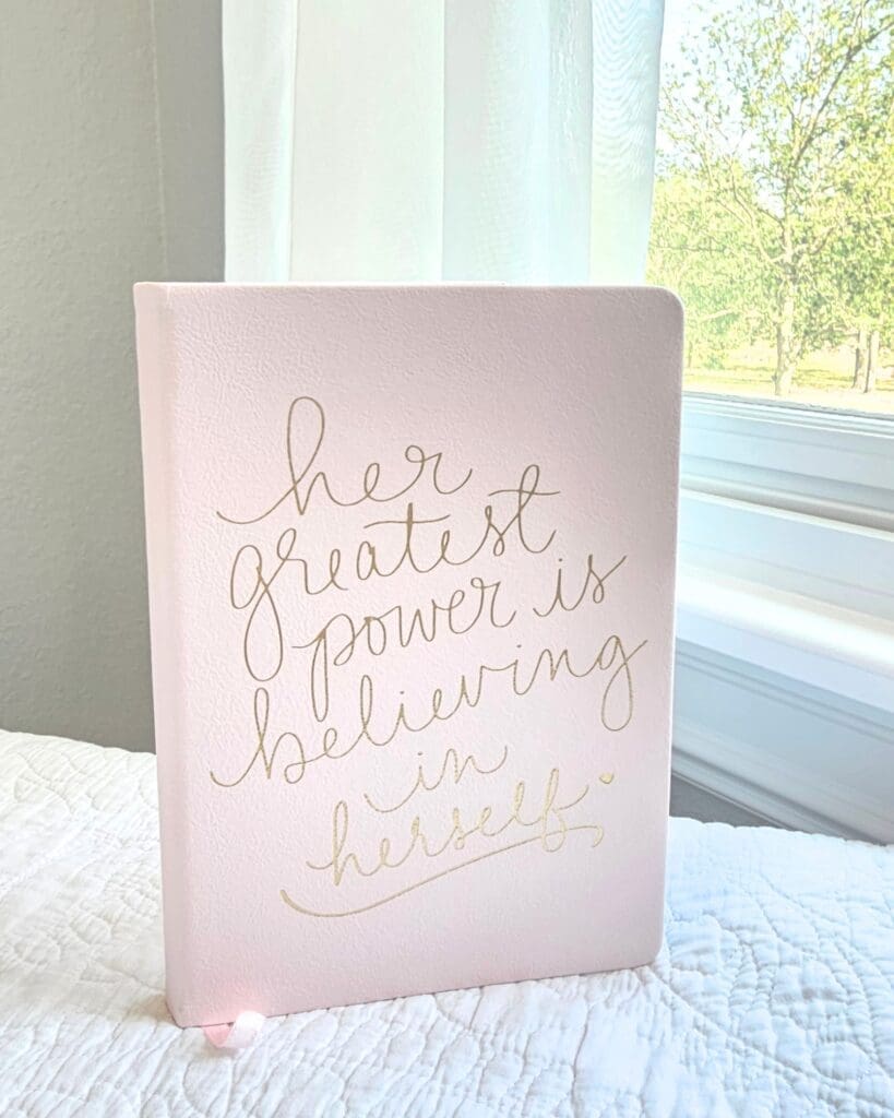 This is a photograph of a small pink notebook with "her greatest power is believing in herself" written in gold cursive font on the cover.