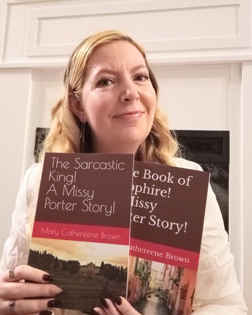 This photograph shows Mary holding two books from her Missy Porter series. Mary has long blond hair and is smiling at the camera. She is wearing a white long-sleeve shirt.